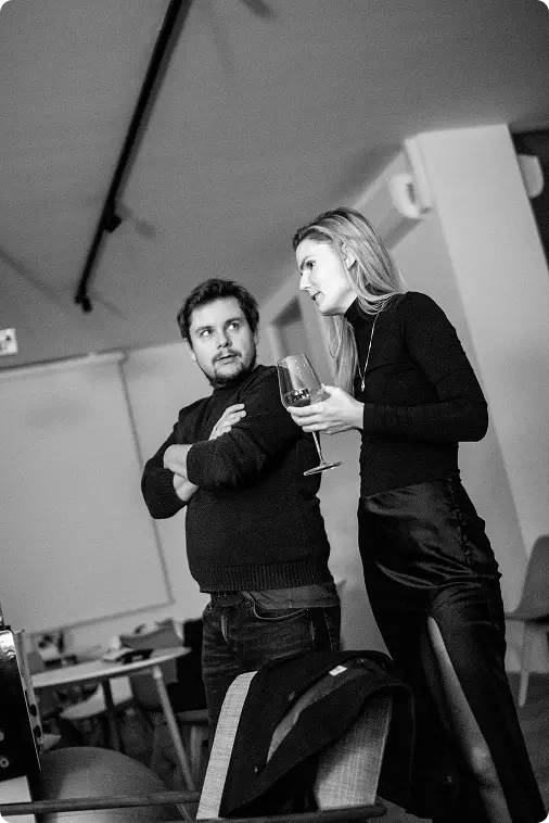 Stephan und Susanne (schwarz/weiß Foto) stehen nebeneinander im Büro. Susanne hält ein Glas in der Hand, Stephan sieht zu ihr herüber